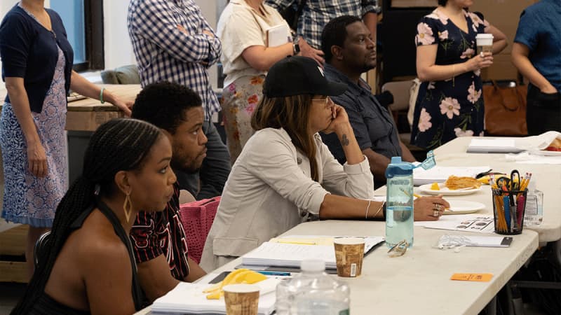 The company of THE REFUGE PLAYS sit at and stand behind long white tables with scripts, beverages, and pencil cans on them.