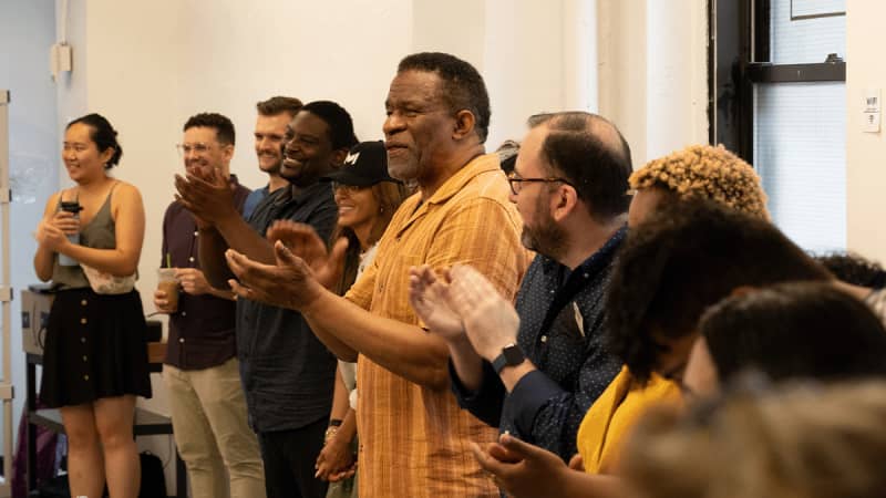 The company of THE REFUGE PLAYS stands in a circle around the rehearsal studio and smiles while clapping.