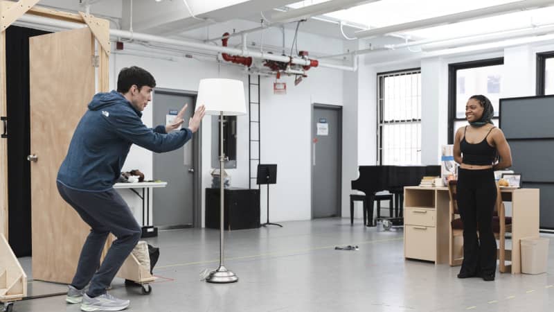  A young man stands by an open door and excitedly gestures to a smiling, reserved young woman standing by a desk on the other side of the rehearsal room.