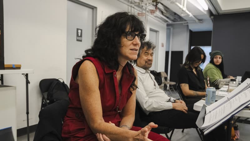 A white woman with wavy black hair and bangs, wearing glasses and a red jumpsuit, sits behind a music stand in a rehearsal room and speaks to the room.