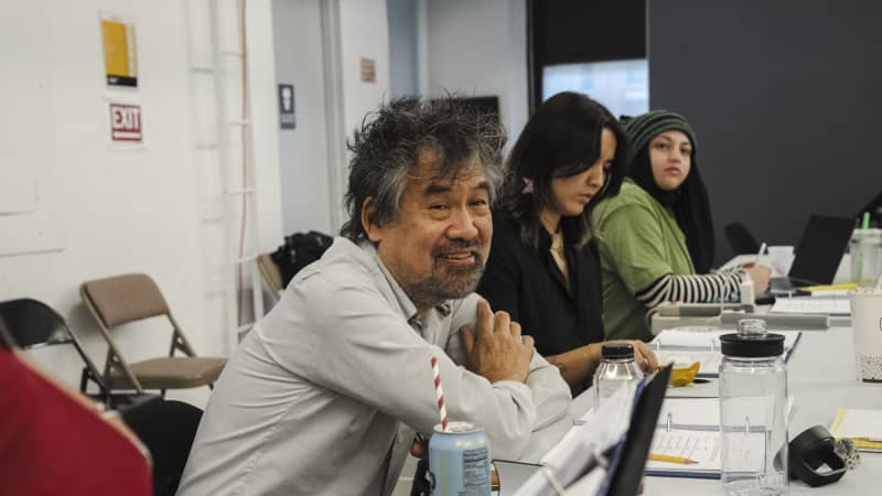 An Asian man with messy dark gray hair and facial hair smiles and leans on a rehearsal table with a script in front of him. 