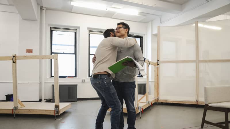 A white man hugs a smiling Asian man wearing glasses and holding a script in the rehearsal studio