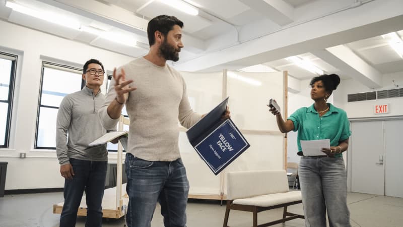 In a rehearsal stuido, a white man holds a script entitled "YELLOW FACE" and stands between an Asian man in glasses and a Black woman holding a tape recorder out to him.