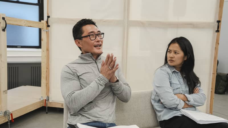 An Asian man wearing glasses sits on a rehearsal bench and clasps his hands while smiling. An Asian woman next to him crosses her arms and glares at him.