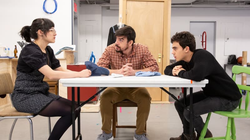 In a rehearsal studio, three actors portraying family members sit around a kitchen table in a tense discussion.