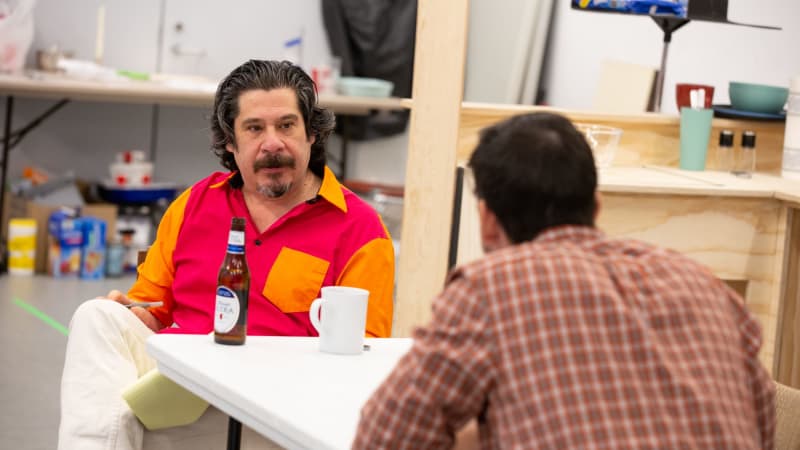 An older man with longer, graying hair and graying facial hair sits across from a younger man in a flannel. A beer bottle and coffe mug sits on the table between them.