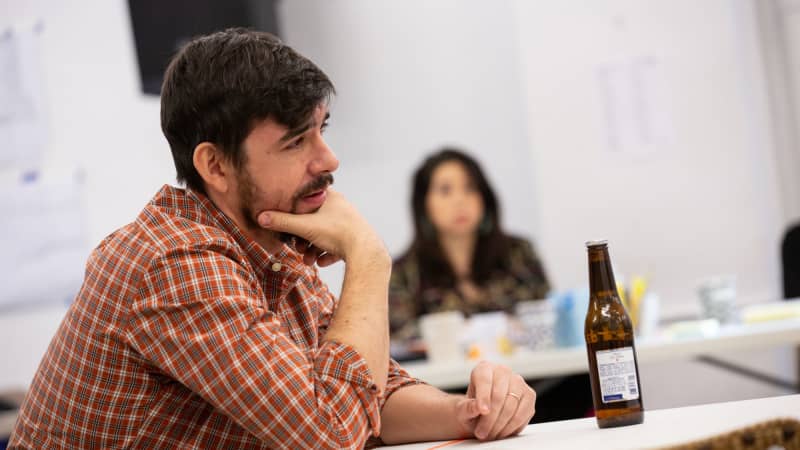 A man in a flannel with short dark brown hair and a light beard rests his face in his hand while leaning on a table with a beer bottle.