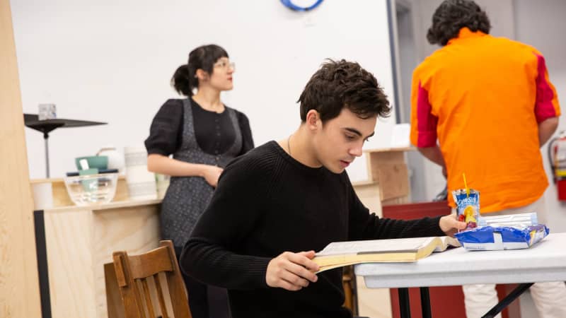 A young man sits at a table in a rehearsal kitchen set and flips through a thick book.