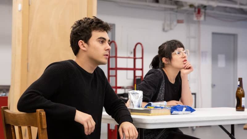 A young man and woman sit at a kitchen table in the rehearsal studio. On their table is a pack of cookies, a Capri Sun, and a beer bottle.