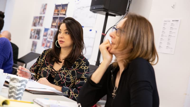 Two women sit behind white tables in a rehearsal studio. In front of them are scripts in binders, and behind them are set and costume inspiration photos.