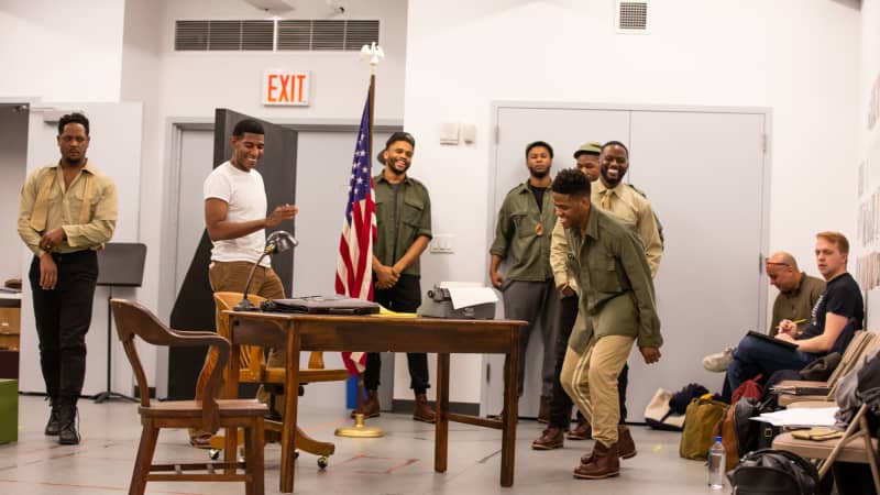 A group of Black men dressed as soldiers stand near the sides of the rehearsal space, not in a scene, and smile and laugh with the whole room.