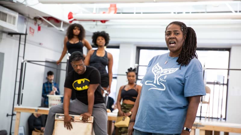 A Black woman with her hair in a side ponytail sings in the rehearsal room while a group of actors stand behind her on a staircase and a washing machine.