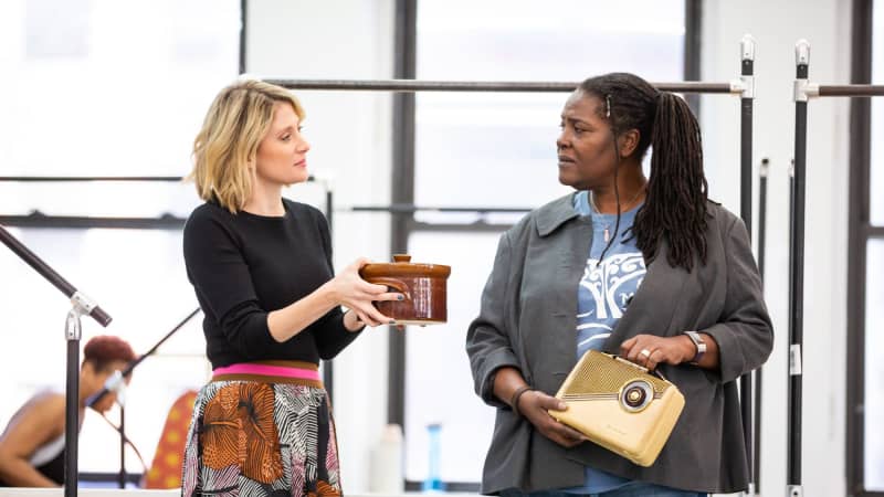 A white woman holds a wooden container to a Black woman holding an old radio in the rehearsal room.