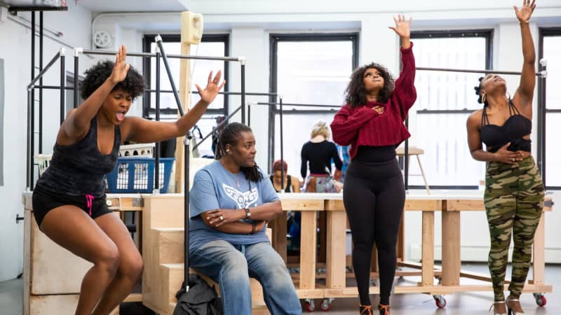 Three Black woman passionately sing with arms raised around another woman sitting on a step with her arms crossed.