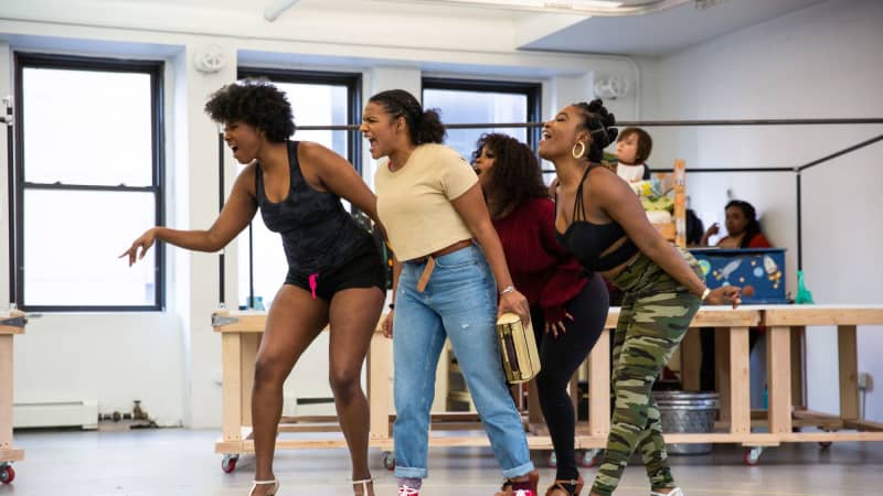 A group of Black women sing in the rehearsal studio. Three of them wear high heels while a younger one wears red Converse sneakers.