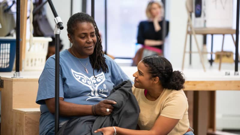 A Black woman sits on steps while a younger Black woman crouches below her. They both smile warmly.