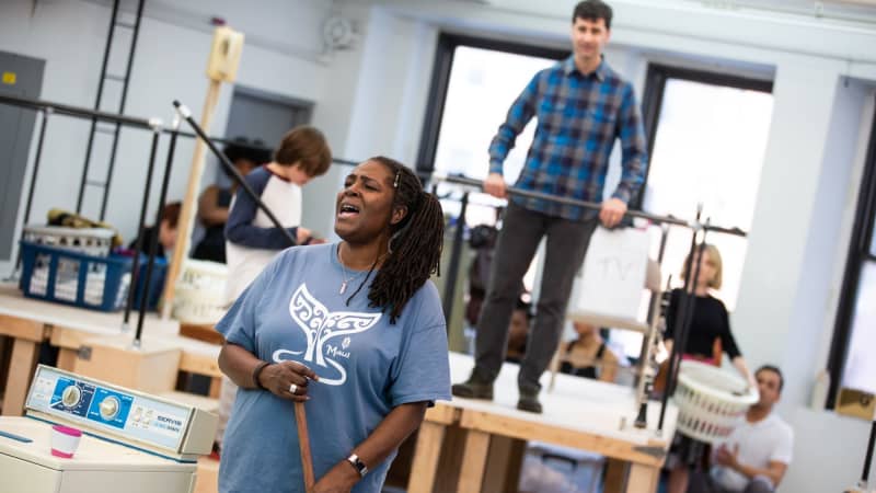 A Black woman holding a broom sings next to a washing machine while a white man stands on a platform behind her and watches.