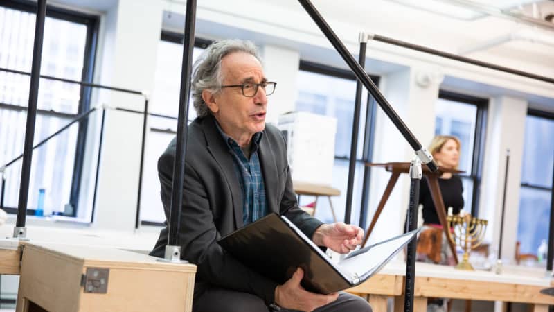 An older white man sits on steps in the rehearsal room and holds a script in a binder.