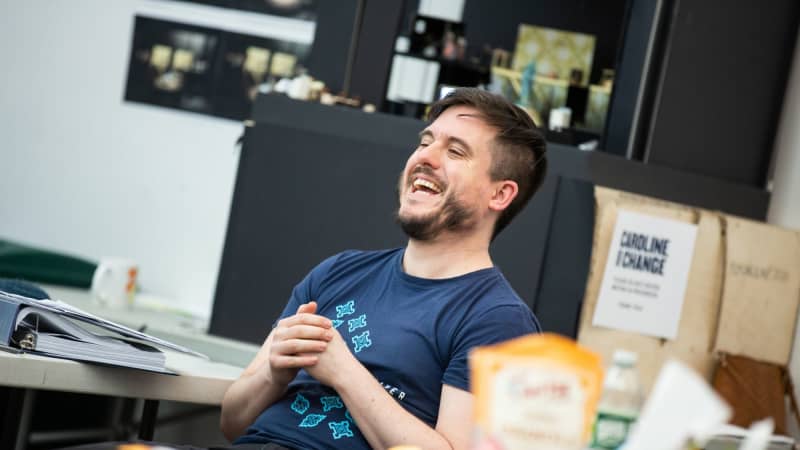 A white man with brown hair and a scruffy beard laughs behind a table in the rehearsal room.