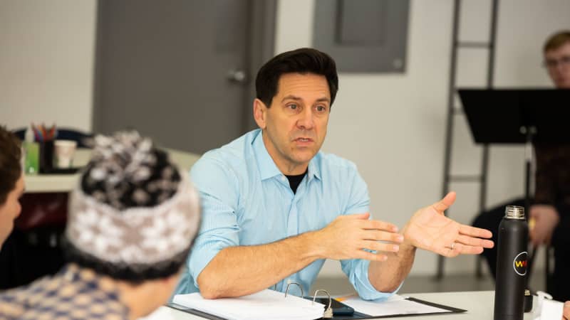 A man at a rehearsal table leans his elbows on his script and gestures his hands while he talks to a group seated around the table.