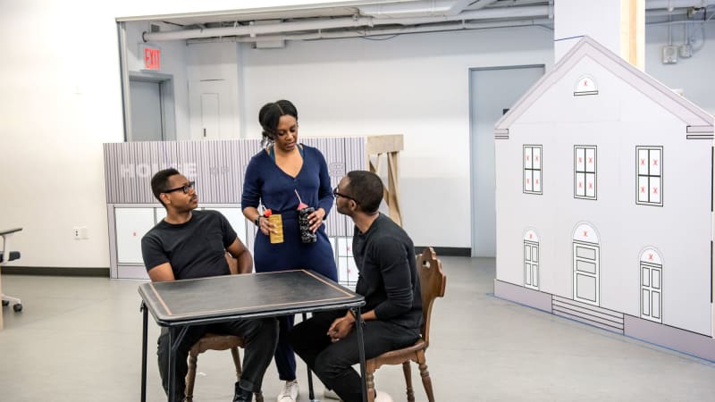 Three dark skinned actors, two male and one female in the middle, sitting at a table in a bright rehearsal studio. There are small building mockups in the background. 