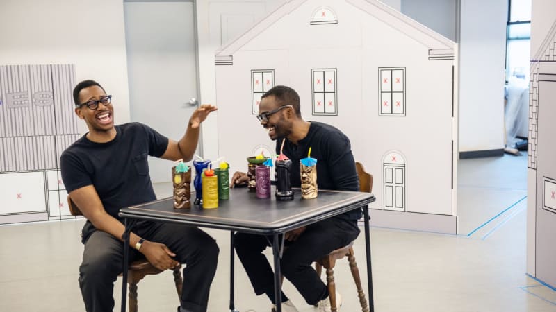 Two dark skinned men sitting at table laughing in a bright room with small scale buildings behind them.