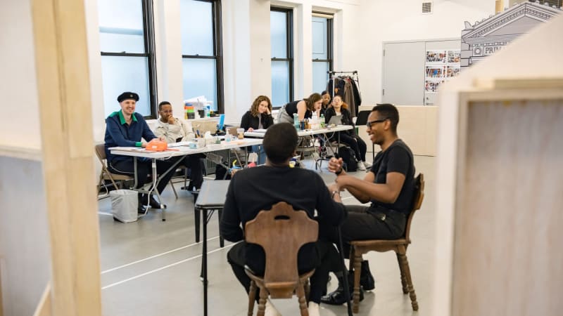 Two dark skinned men sitting at a table with their backs towards the camera. The creative team sitting at a long table in front of windows watch the two men in the scene.