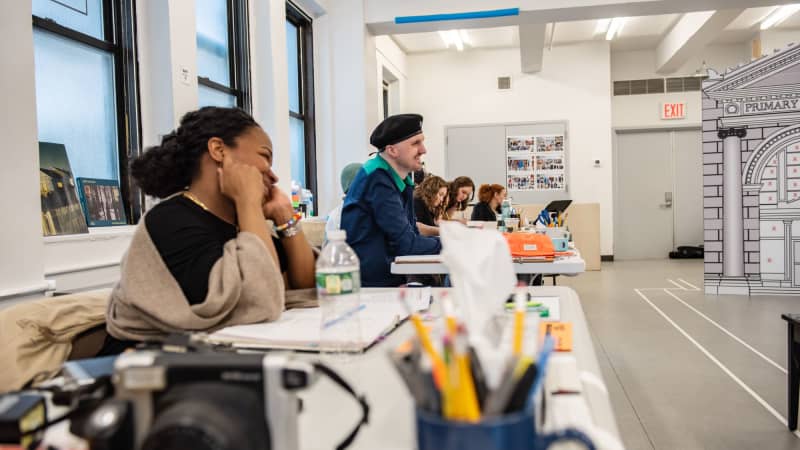 A medium-dark skinned woman and a light skinned man sitting at a white folding table in a bright rehearsal studio with folders and other office supplies. 