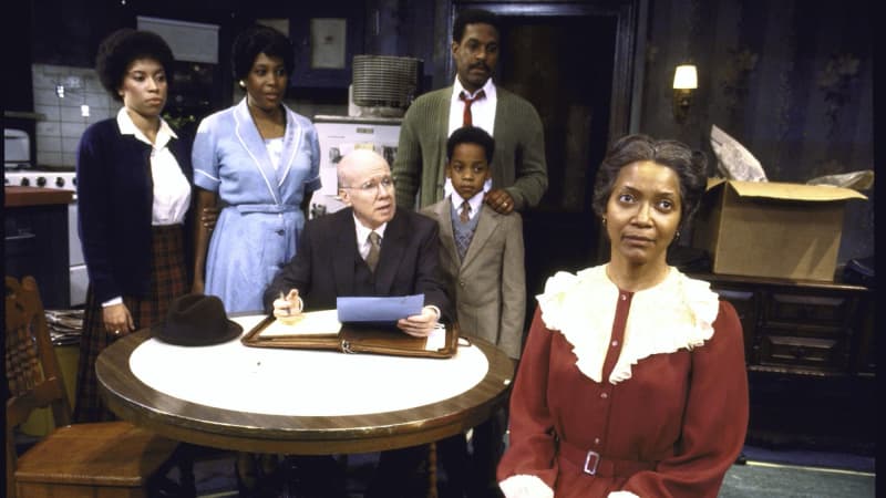 The cast of "Raisin in the Sun" on stage. 5 actors are behind a table, one sitting, while a woman in a red dress sits upstage towards the camera.