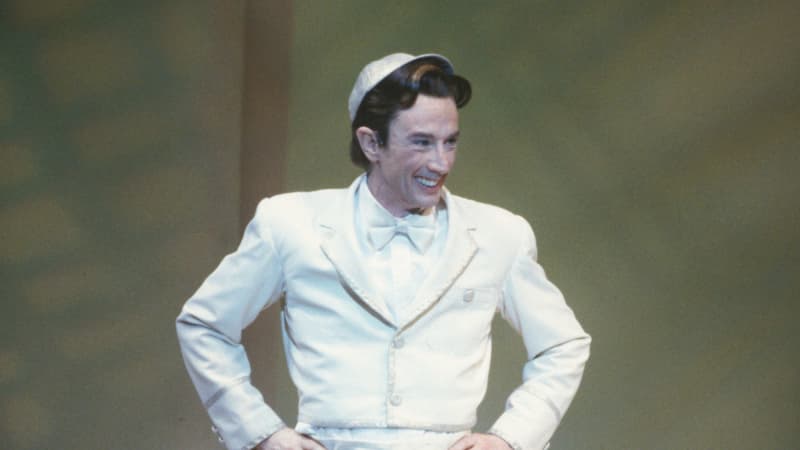 Martin Short in a small white suit and white cap. His hands are on his hips and he is smiling. He stands in front a blurred green background.