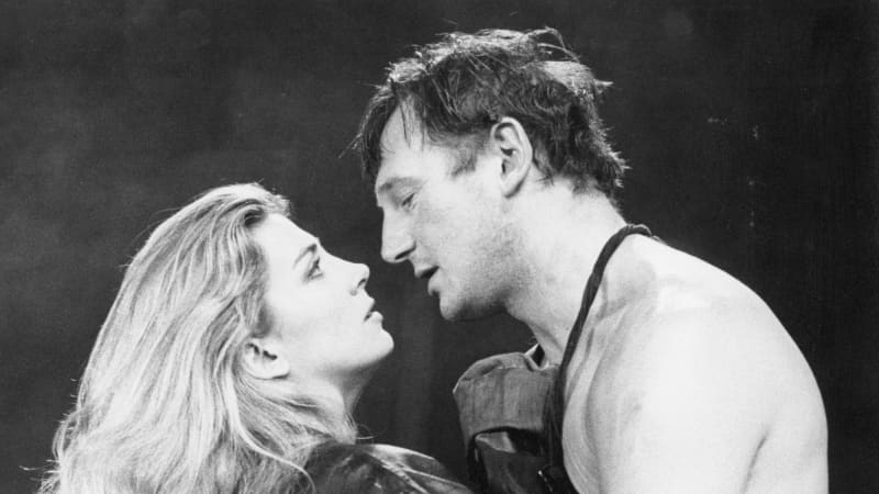 Black and white photo of Natasha Richardson and Liam Neeson embracing one another. Liam (right) is wearing an apron while Natasha (left) is wearing a black leather coat. 