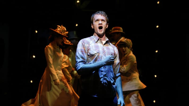 Neil Patrick Harris in a flannel shirt  and a spotlight in front of several actors in two lines in dark light.