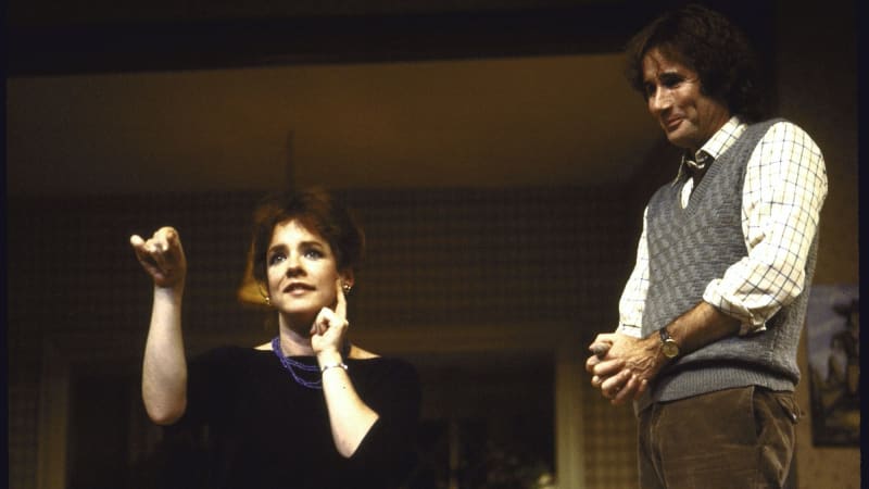 Stockard Channing and Jim Dale in A Day in the Death of Joe Egg. Stockard Channing is wearing a black dress and has one hand to her cheek and the other in the air. Jim Dale is in a gray sweater vest w