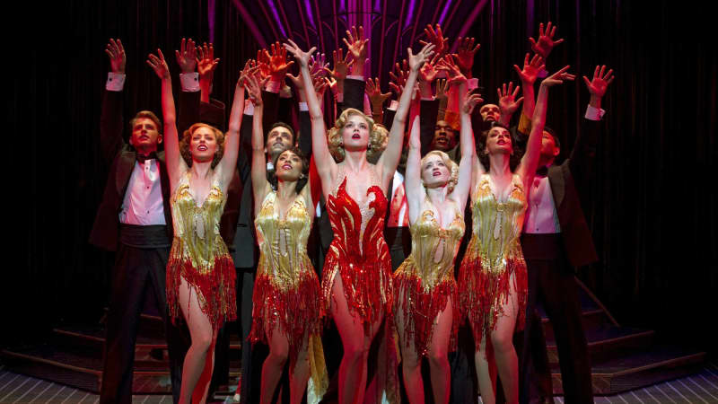 "The cast of Anything Goes; in red and gold costumes on stage. They are gathered closely together and look upwards with their arms raised and palms facing forward."