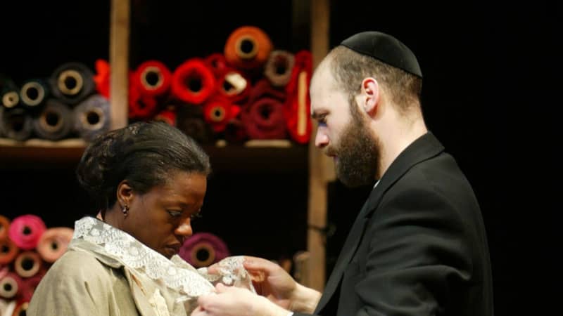 Viola Davis (left) in a white dress being buttons up by Corey Stall (right) in a black coat. A shelf of rolled up fabric sits behind them.