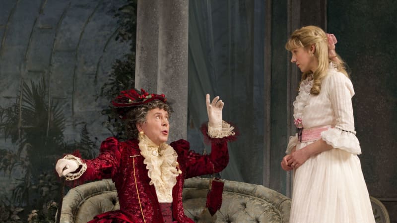 Two actors on stage, the older one sitting (left) on a couch raising their hands to a younger light skinned actor (right) who is wearing a white dress with pink accents.