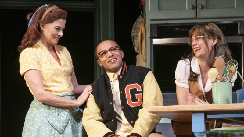 A young smiling Black man in a letterman jacket and wearing glasses sits in a kitchen with two white women on either side of him. One of them rests a hand on his shoulder.