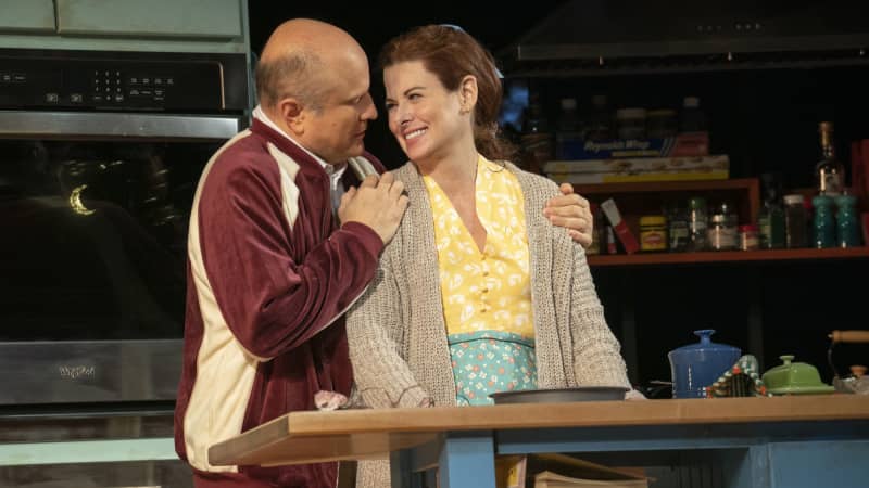 An older man puts his arms on the shoulders of a woman wearing a yellow dress and an apron. They smile at each other while standing in a kitchen.