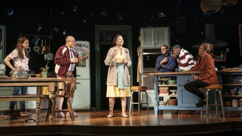 A group of smiling actors sit and stand around a kitchen-themed set on stage.