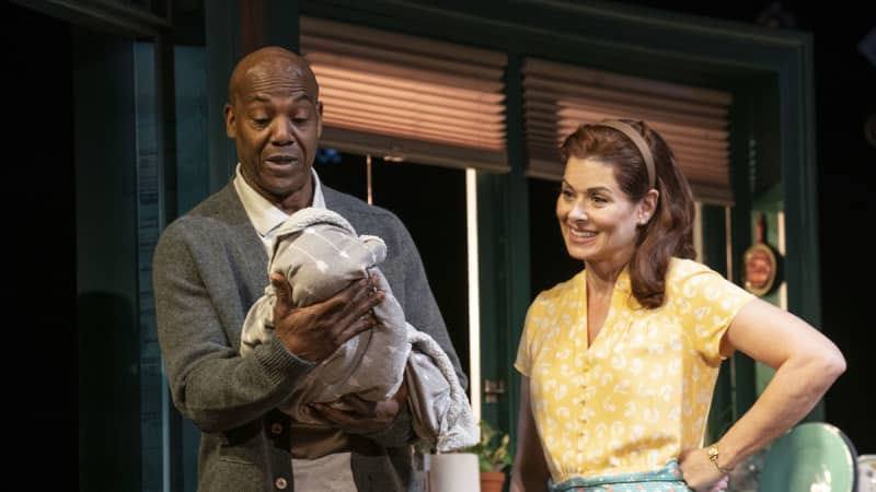 A Black man holds a blanket with a baby in it while a woman in an apron stands next to him, smiling at the baby.