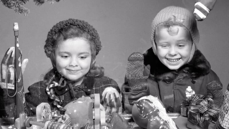 Two small, light-skinned children in winter hats smile at a display of toys.