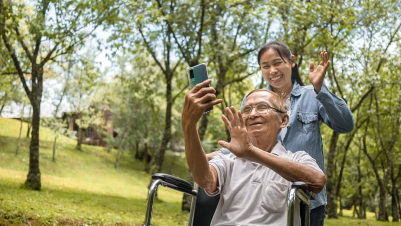 A medium-light skinned older man in glasses, using a wheelchair, holds a smartphone out and waves to the person on the other end of the call. A young medium-light skinned woman stands behind the wheel