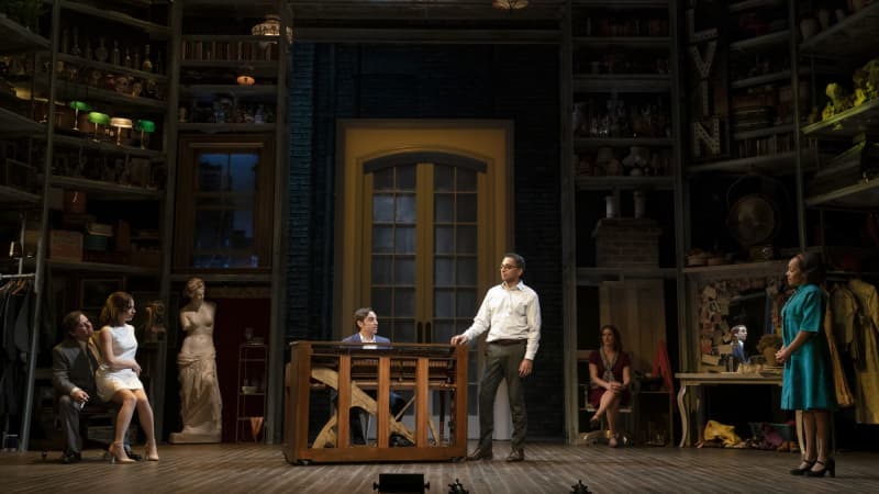 An octagonal room. Tall shelves along the walls. There are tall doors up center, and a Greek sculpture of a woman left. A light-skinned man plays piano as a medium-skinned man sings. Others look on.