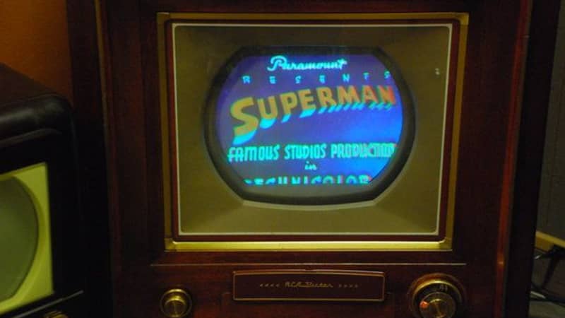 A small, oval screen set inside a dark wooden case. The screen displays a blue background with the word ‘Superman’ over it in reddish-orange letters. 