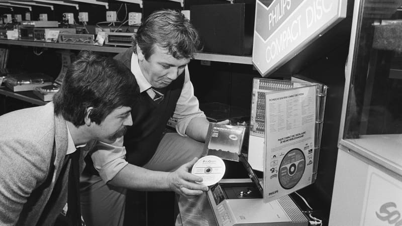 A light-skinned man in an oxford shirt, tie, and sweater vest holds a compact disc as a light-skinned man with a dark mustache leans forward to look at it. 