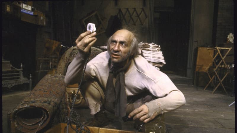 A light-skinned, unkempt man in a shirt with holes in the cuffs squats in front of a wooden box full of cords. Behind him, a stack of newspapers and a rolled-up carpet. 