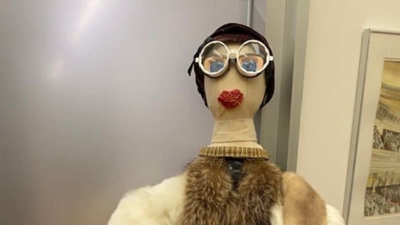 A stuffed muslin head atop a puppet body. The face has yarn lips and blue, sewn eyes. The figure wears draped furs, large-framed glasses, and a black hat.