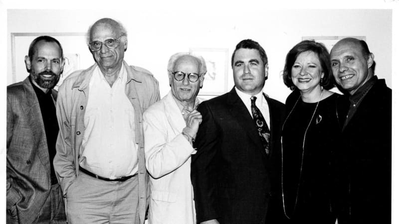 From left, four smiling, light-skinned men in suits, then a smiling, light-skinned woman in a dark dress and long necklace, then a smiling, light-skinned man in a sport coat.