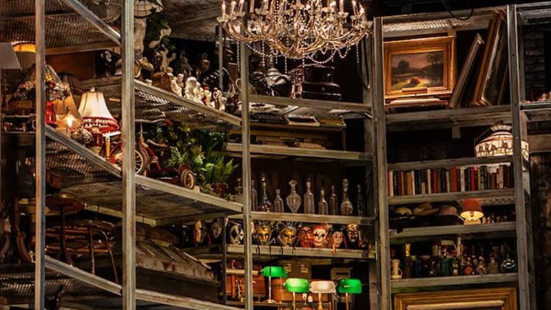 Large metal shelves, each organized to hold one type of object: green-shaded lamps, decorative masks, clear glass bottles, framed artwork. A chandelier hangs in front of the shelves.