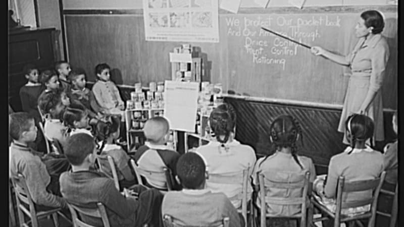 A dark-skinned woman in a dress uses a pointer to indicate a section of a chalkboard for her students, a group of dark-and-medium-skinned elementary school children.